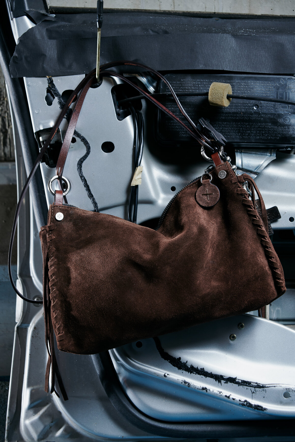 An INA KENT brown suede handbag with leather straps hangs elegantly on the exposed metal frame of a car door.