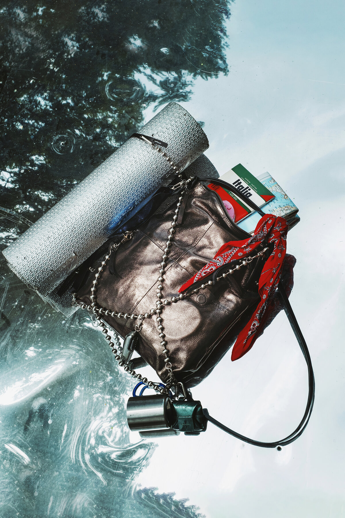 A metallic mesh INA KENT bag with a chain strap, red bandana, silver mat, Italian cigarette rolling papers, and metal padlock sits on a reflective surface outdoors.