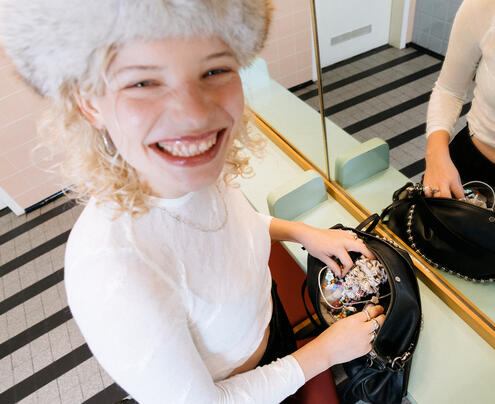 Luisa Gaffron with a beaming smile in a pastel-colored room. She is wearing a white blouse and a fluffy hat while she searches for something in her INA KENT bag - between colorful beads and a scrunchie.