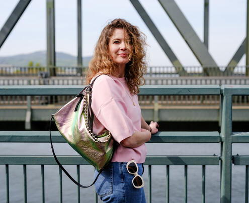 A woman with curly hair in a pink top and jeans stands by a metal railing on a bridge, carrying an iridescent INA KENT bag and holding sunglasses.