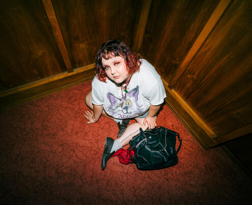 Person with short red hair sits on a carpeted floor in the corner of a wood-paneled room, wearing a cat graphic t-shirt and boots, with an INA KENT black bag beside them.