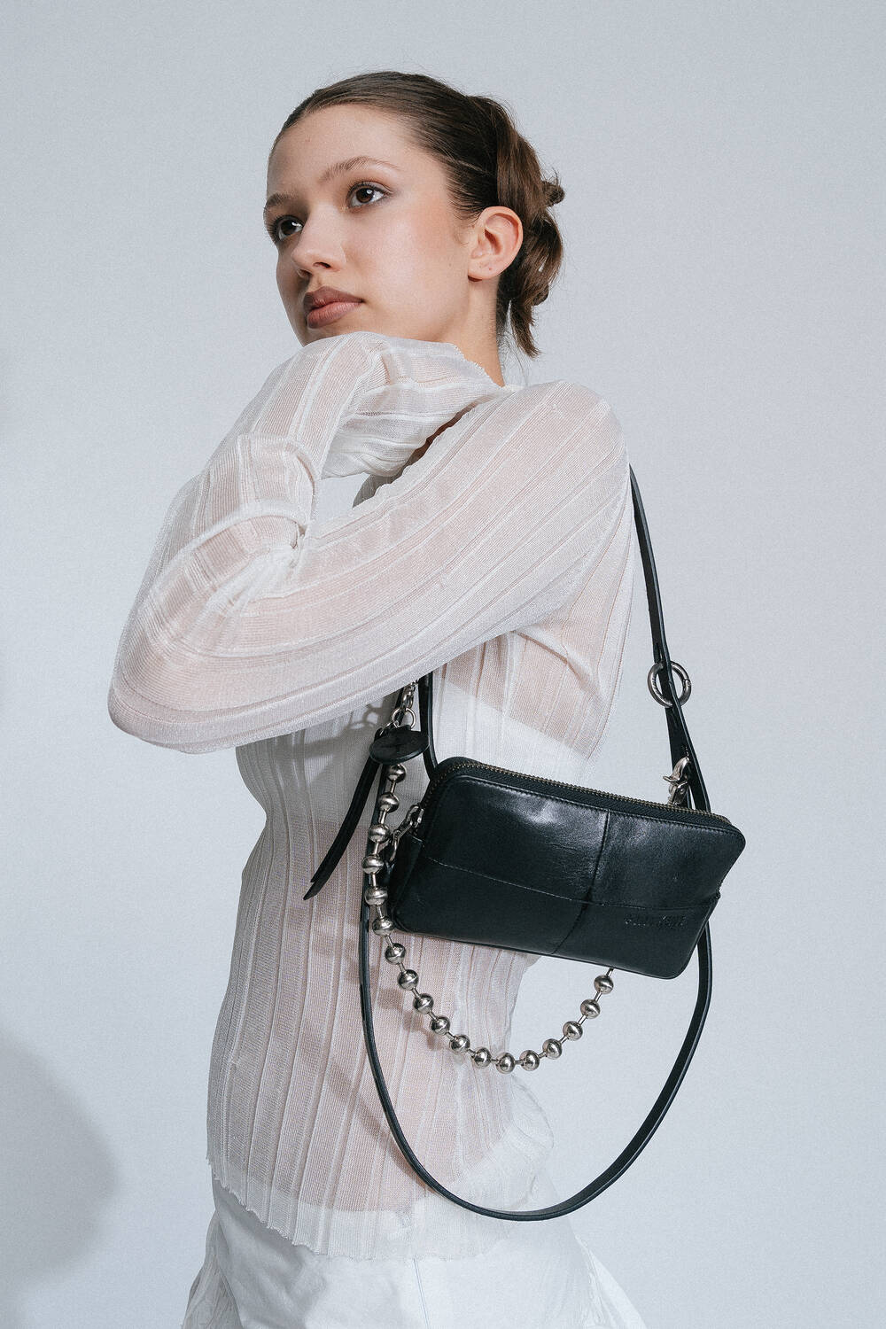 A woman with tied-back hair, wearing a white semi-transparent blouse and holding an INA KENT black rectangular purse with a chain strap, poses against a plain background.