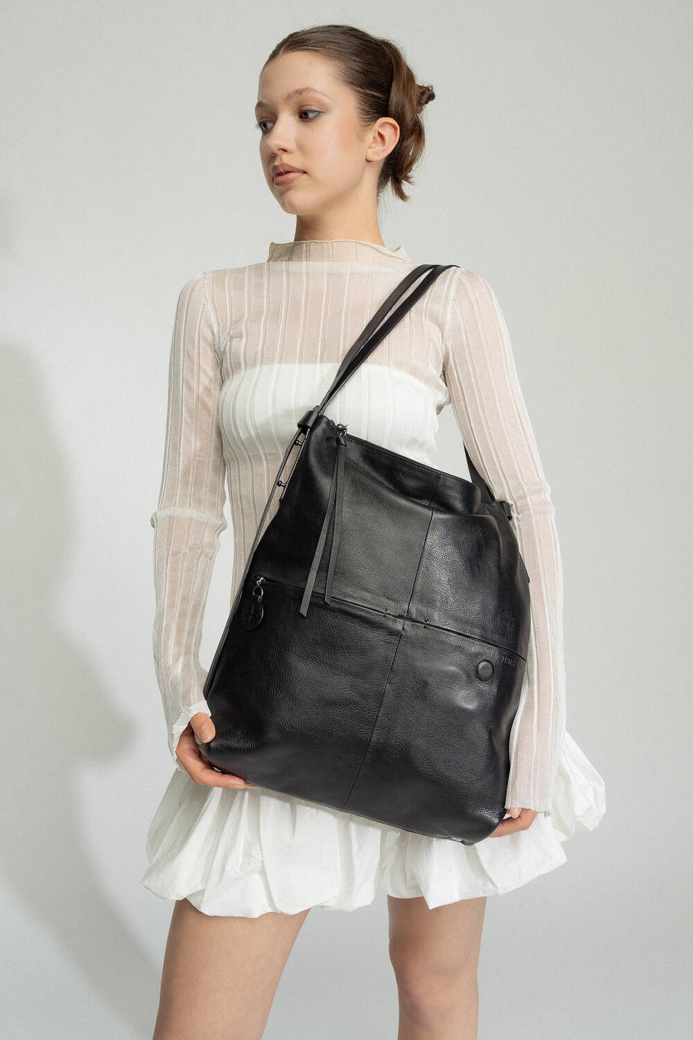 A woman wearing a white sheer top and white skirt holds a large black leather shoulder bag by INA KENT.