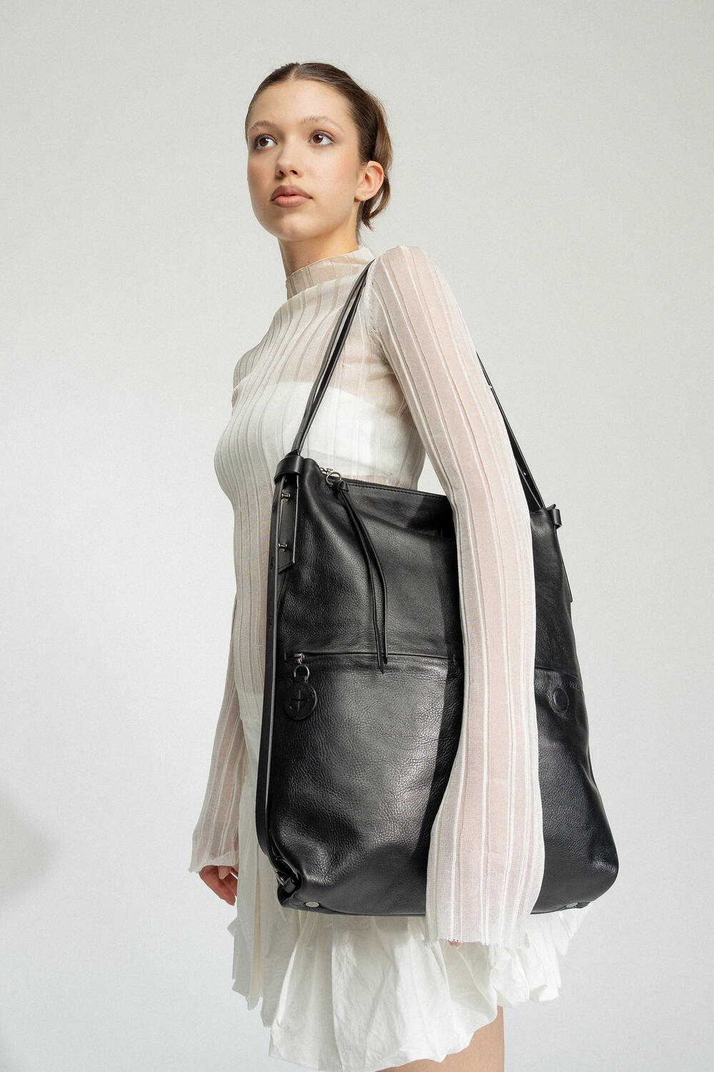 A young woman in a white sheer long-sleeve dress elegantly carries a large black INA KENT leather bag over her shoulder, standing against a plain background.