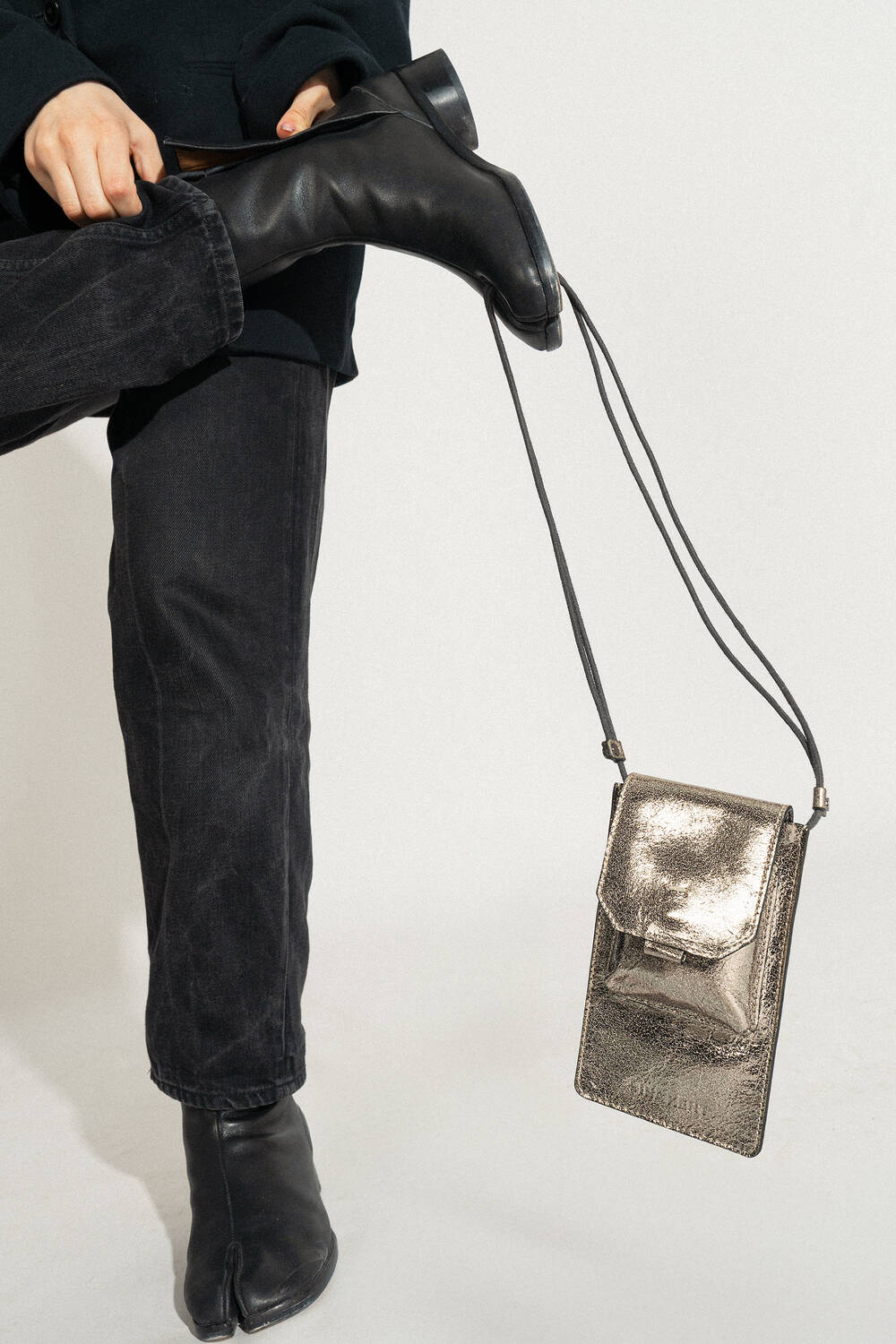Person in black pants and boots lifting one leg, with an INA KENT metallic purse stylishly hanging from their ankle.