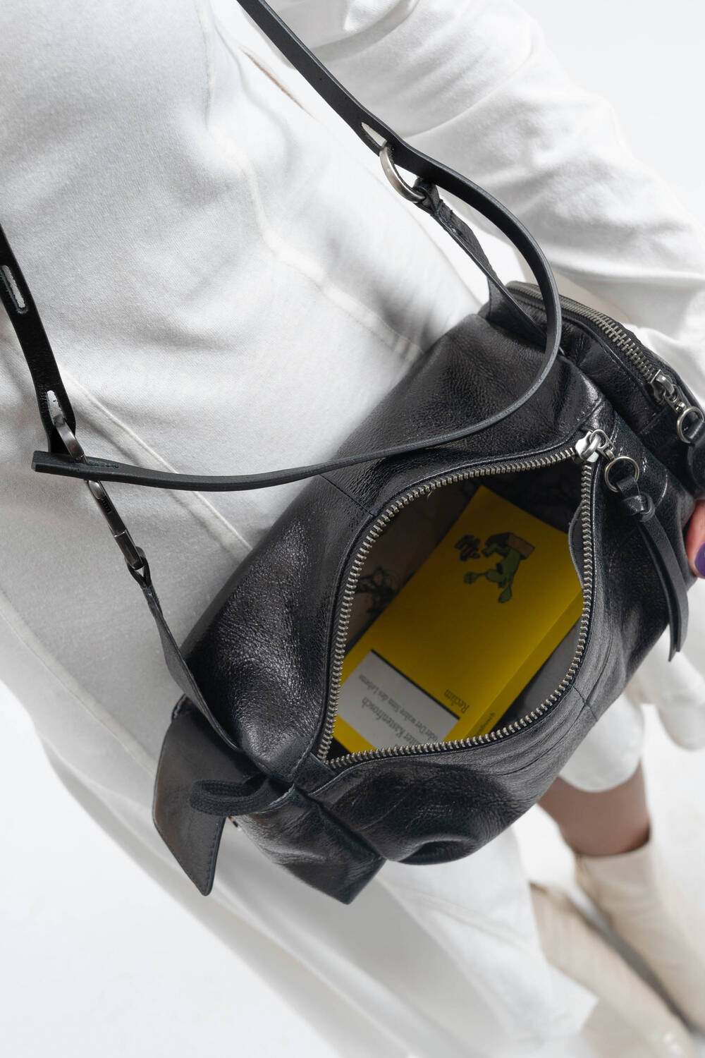 A person in a white outfit carries an INA KENT black leather bag with the zipper open, revealing a yellow book inside.