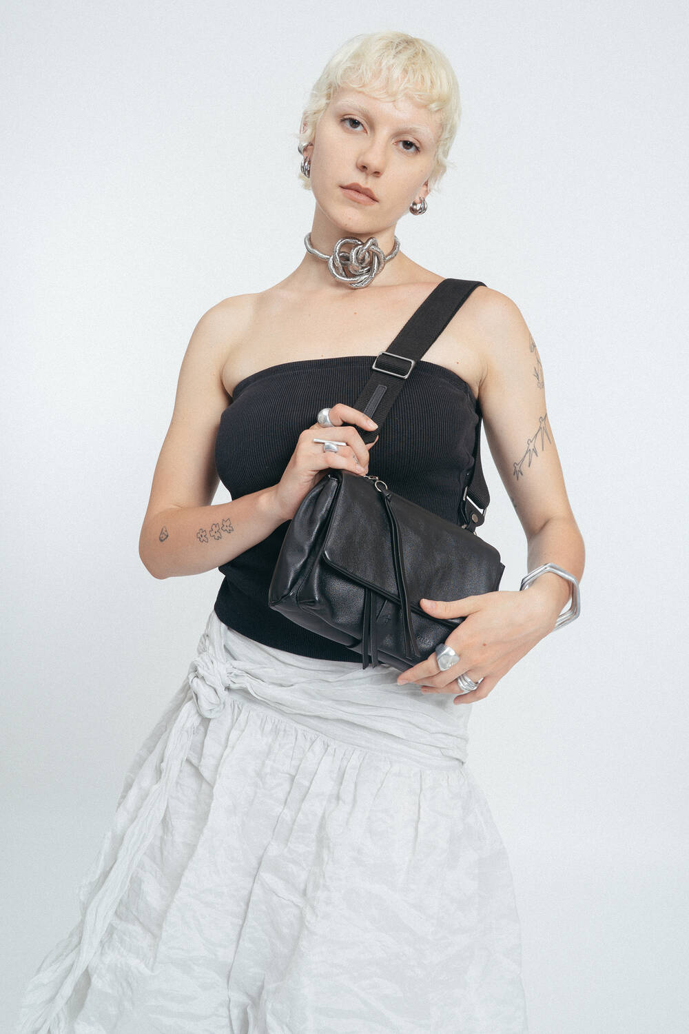A person with short blond hair wears a black strapless top, white skirt, silver jewelry, and holds an INA KENT black shoulder bag against a plain light background.