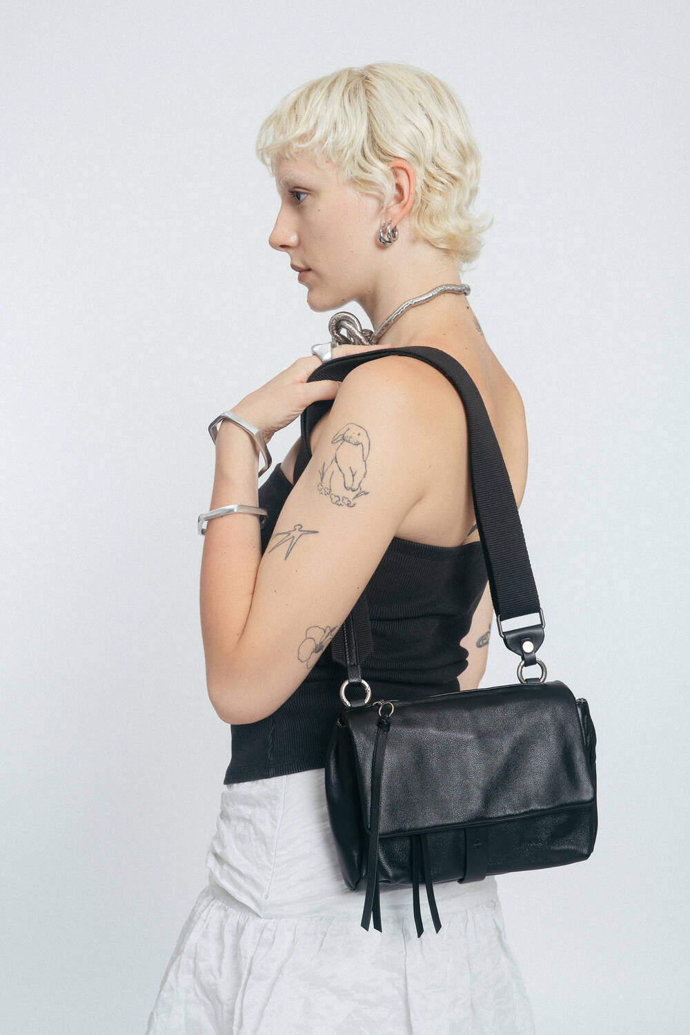 Person with short blonde hair, wearing a black tank top and white skirt, holds an INA KENT black shoulder bag; visible arm tattoos and silver jewelry complete the look.