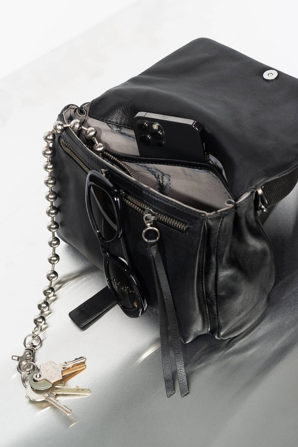 A black leather INA KENT bag with its top open, showing a smartphone, sunglasses, and keys attached to a metal chain on a light reflective surface.