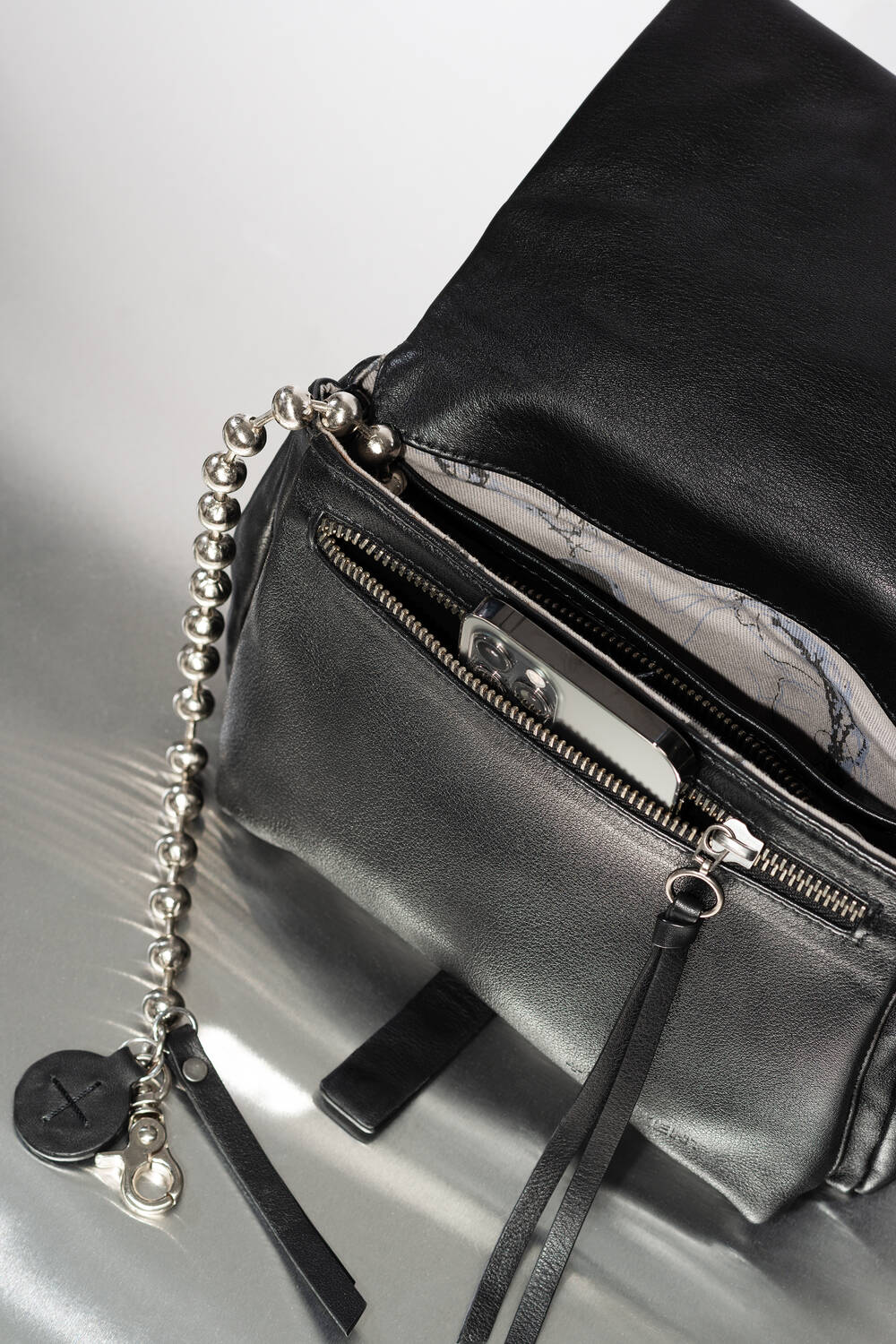A black INA KENT leather bag with a silver chain keychain and a phone partially visible in the front zippered pocket, resting on a reflective surface.