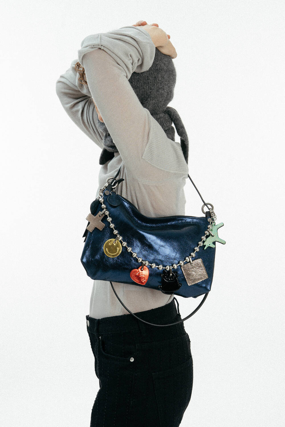 Person in a light long-sleeve shirt and gray headscarf poses with a shiny blue INA KENT handbag decorated with colorful charms over their shoulder.