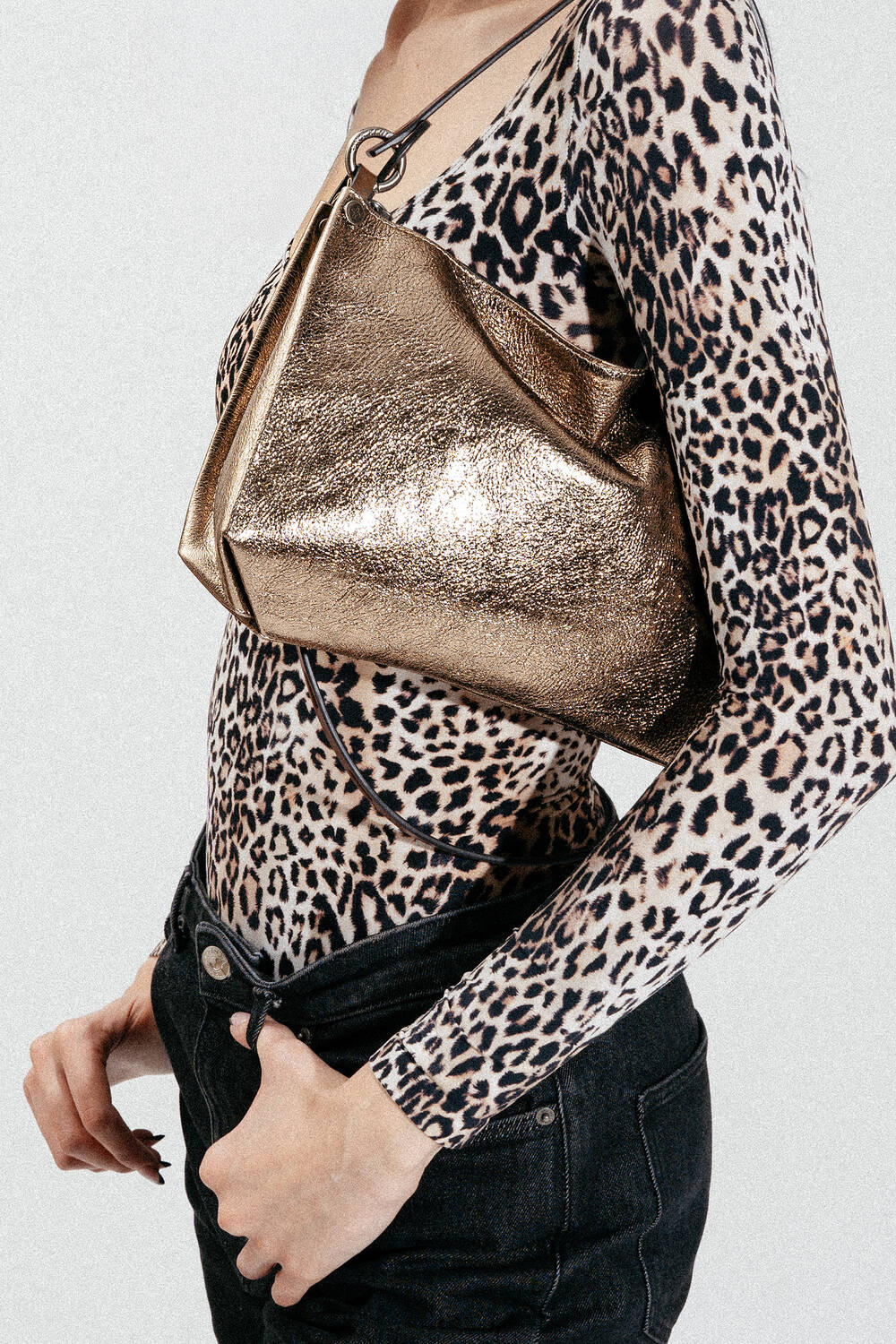 Person, die ein langärmeliges Oberteil mit Leopardenmuster und eine schwarze Jeans trägt und eine goldene Metallic-Handtasche von INA KENT über der Schulter hält.