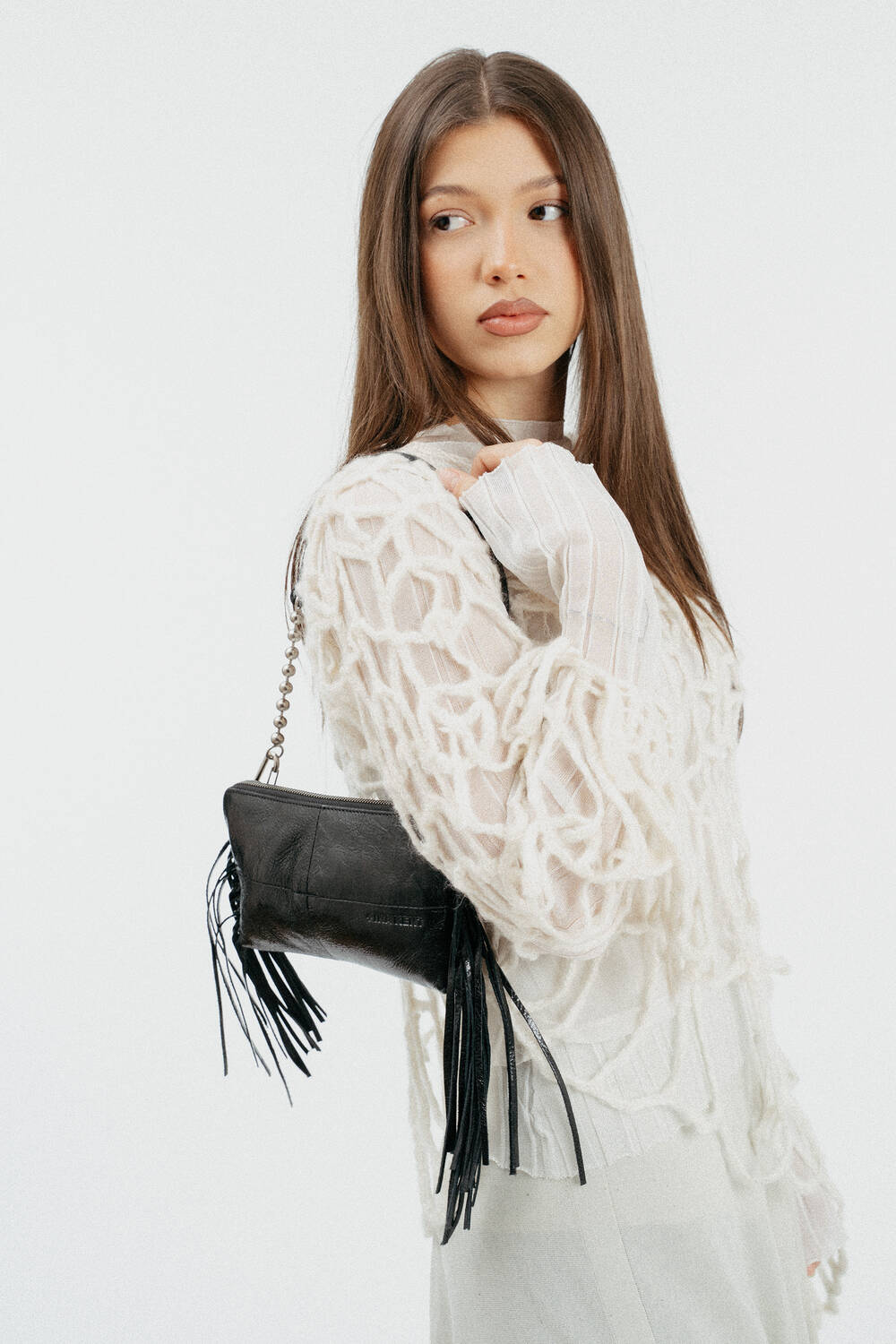 Weiblich gelesenes Modell im losen, weißen Strickpullover und schwarzer, kleinen Ledertasche mit Fransen