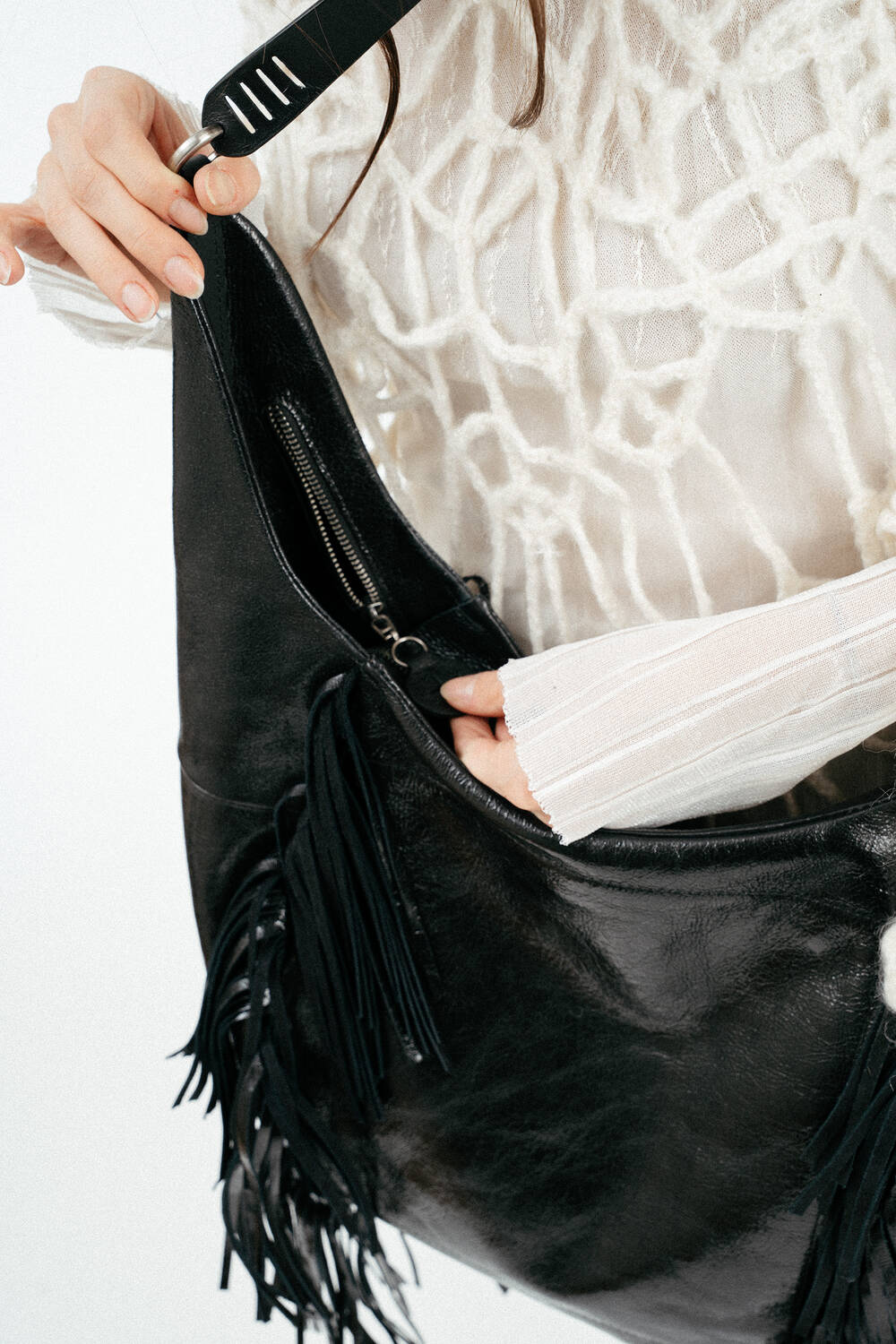 A person wearing a white textured sweater opens a large black INA KENT leather handbag with fringe detailing.