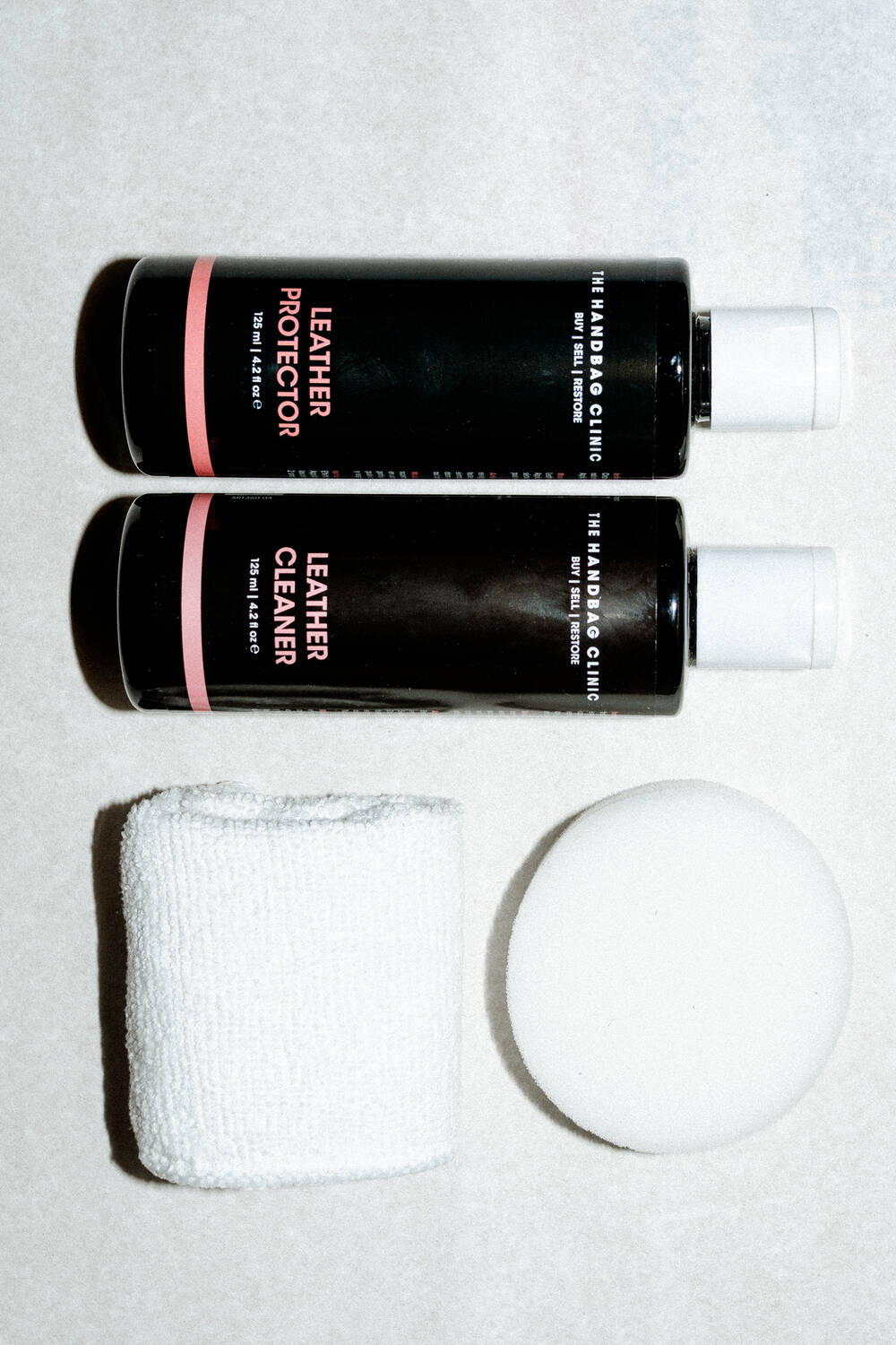 Two black INA KENT bottles labeled "Leather Cleaner" and "Leather Protector" are placed next to a white cloth and a round white sponge on a light surface.
