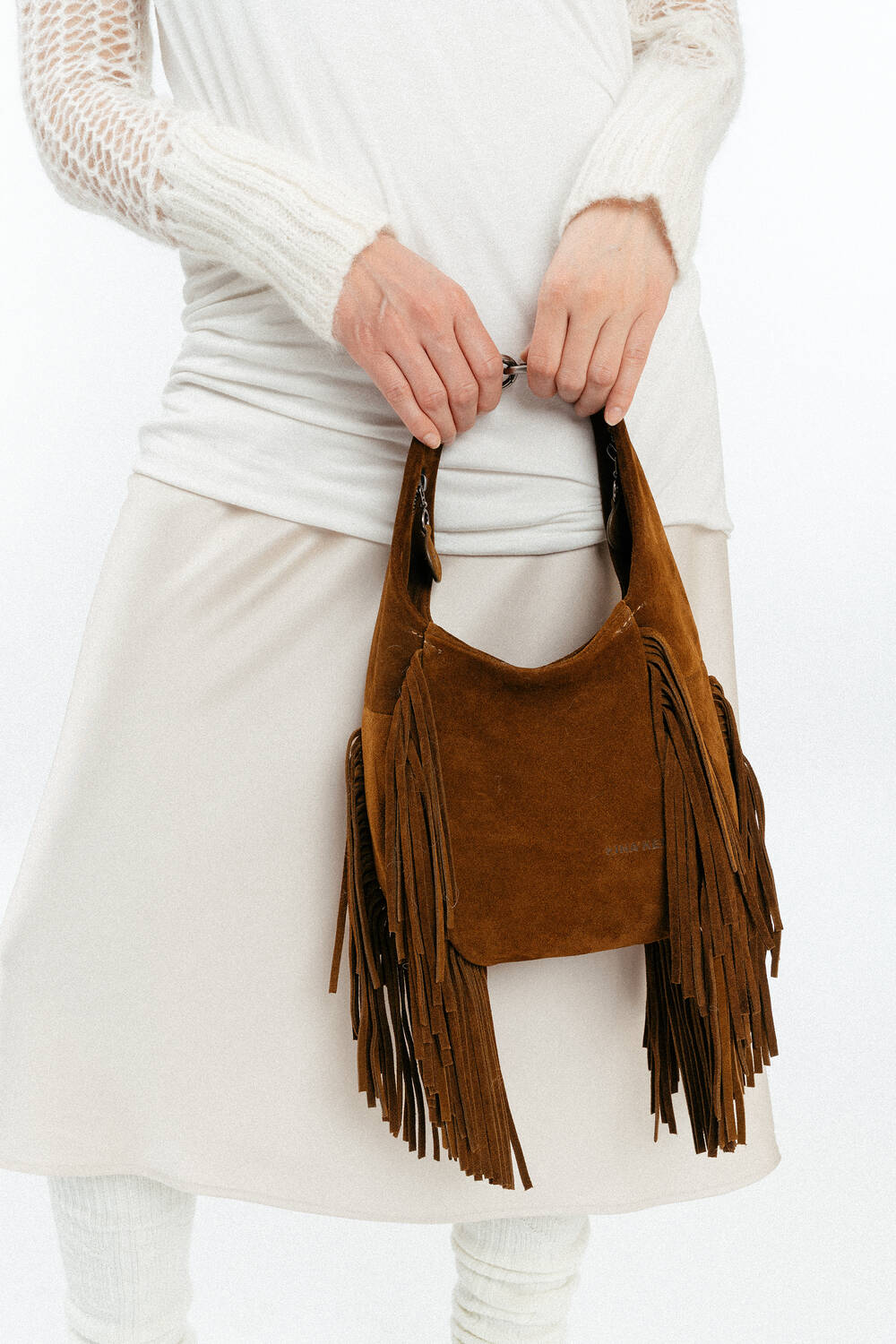 Person holding an INA KENT brown suede fringe handbag, wearing a white long-sleeve top and cream skirt.