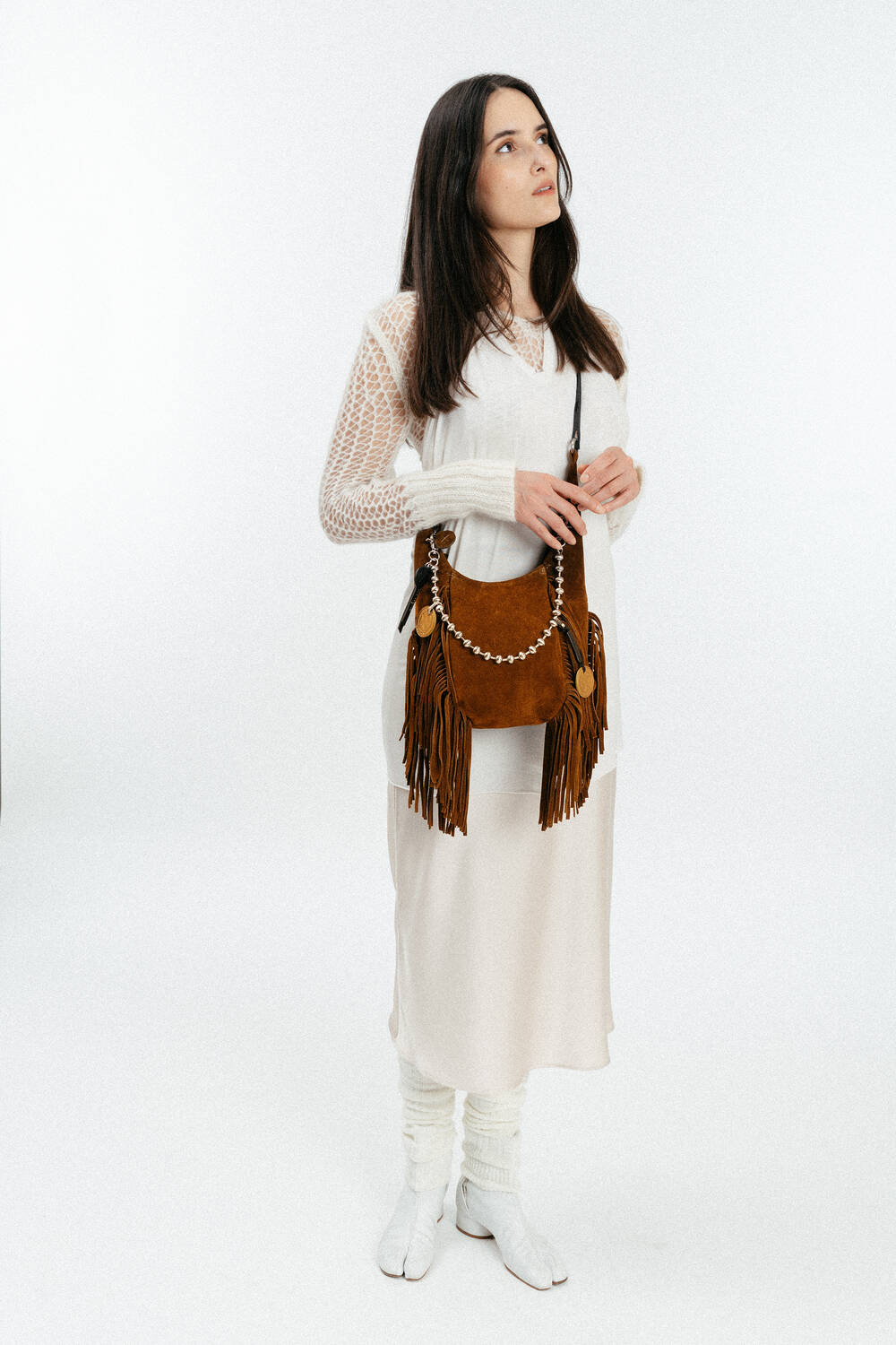 Woman wearing a long white dress, white leg warmers, and white shoes, holding an INA KENT brown fringed handbag, standing against a plain white background.