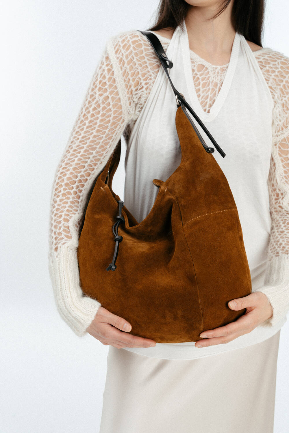 Eine Frau in einem weißen Spitzenoberteil hält eine braune INA KENT-Umhängetasche aus Wildleder mit schwarzem Riemen und Kordelzug.