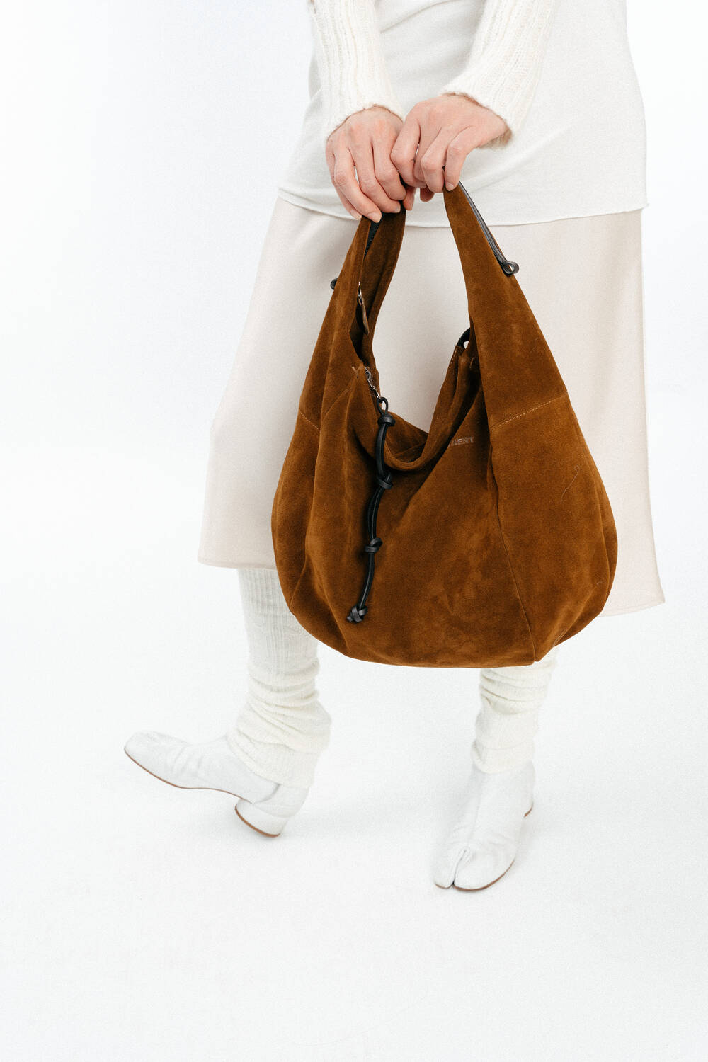 Eine Person in cremefarbenem Outfit und weißen Schuhen hält eine große braune INA KENT-Wildledertasche mit schwarzen Akzenten vor einem weißen Hintergrund.
