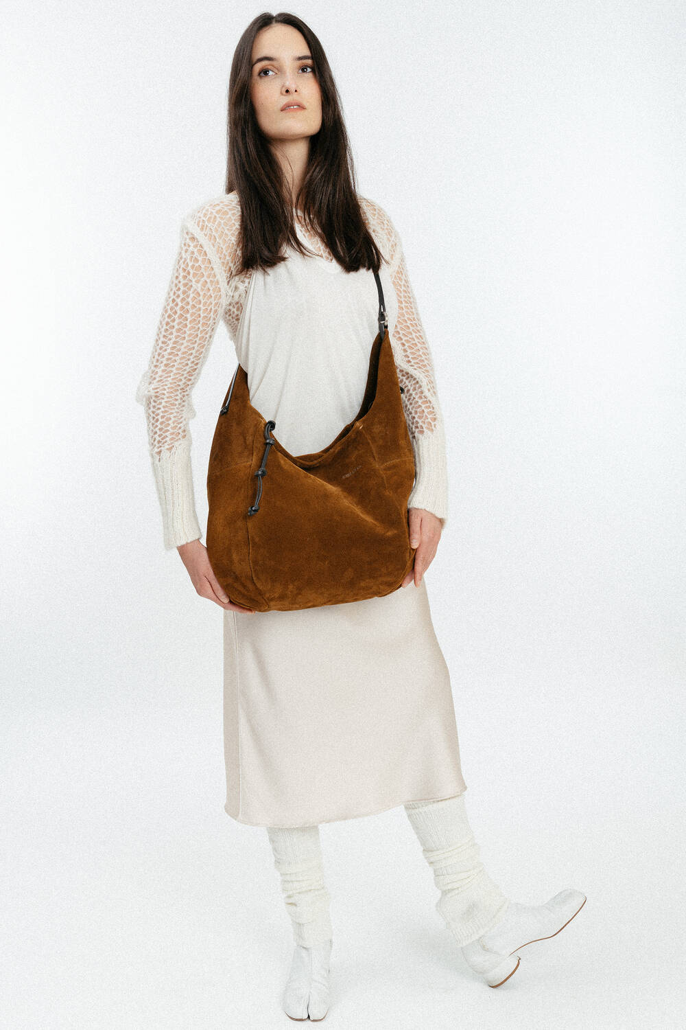Frau in weißem Strickoberteil, cremefarbenem Rock und weißen Stiefeln, die eine große braune INA KENT-Schultertasche aus Wildleder vor einem schlichten weißen Hintergrund hält.