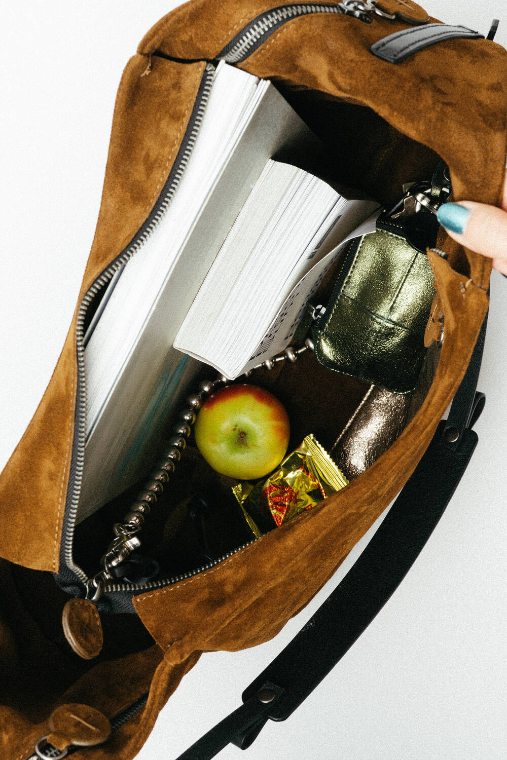 Eine offene braune Wildledertasche von INA KENT mit einem Buch, einem grünen Apfel, Schlüsseln, einer goldenen Brieftasche und einer gelben Bonbontüte.