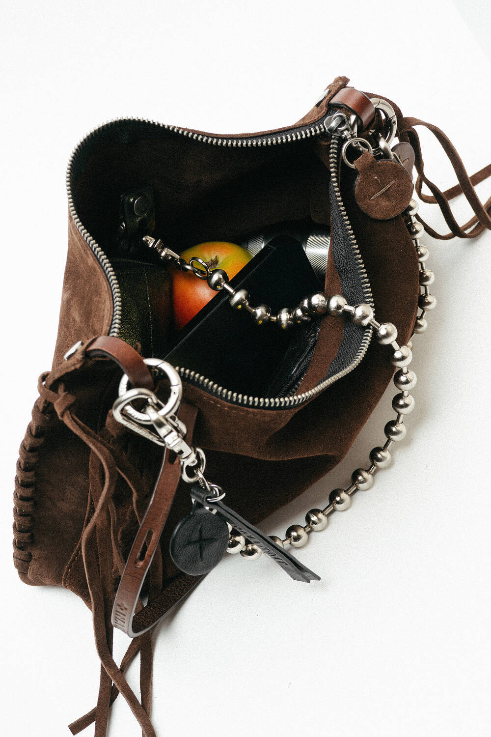 Open brown suede INA KENT bag with a zipper, containing a smartphone, a red and yellow fruit (possibly an apple), and a beaded chain accessory visible inside.