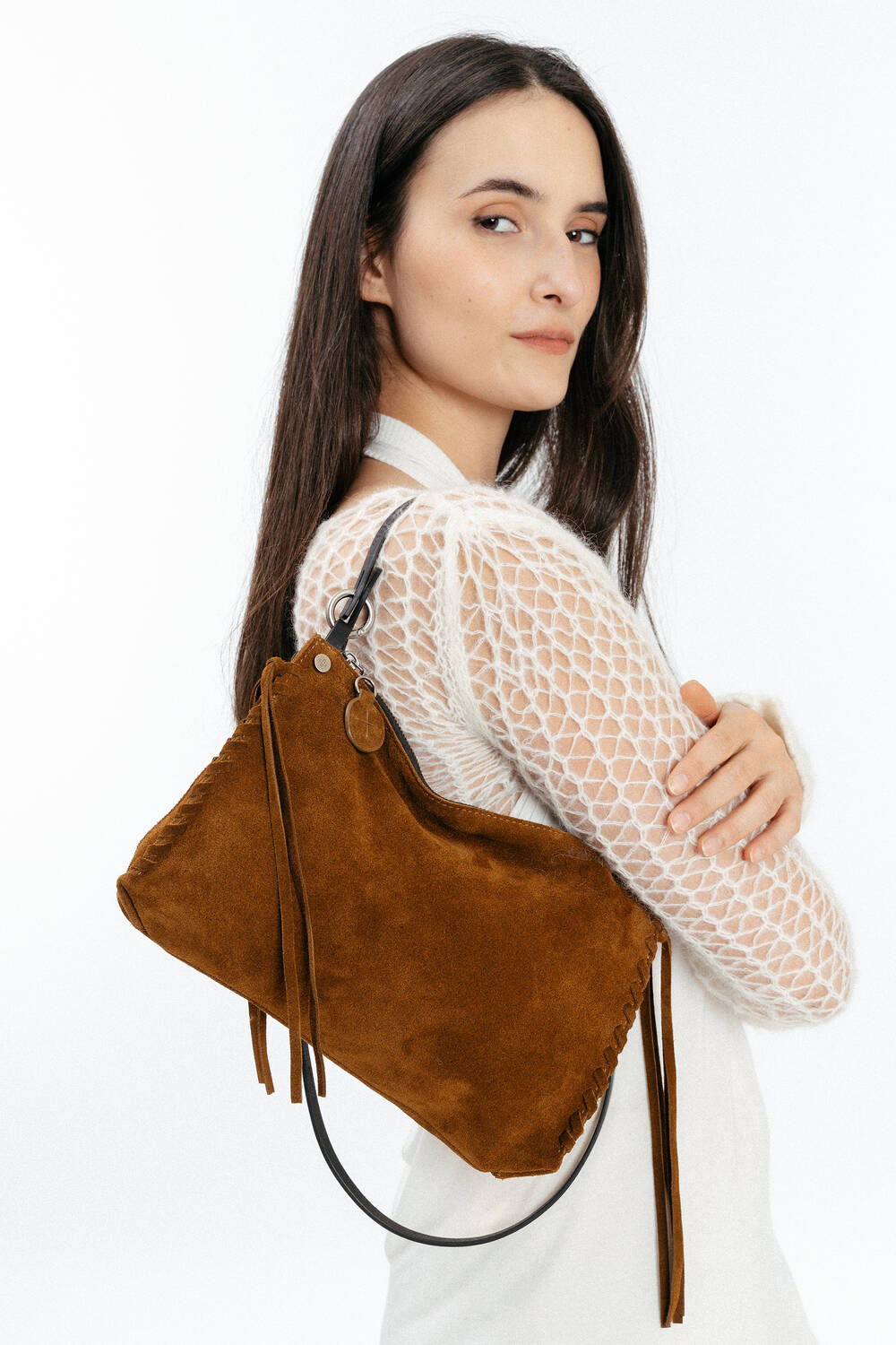 Woman wearing a white textured top holds an INA KENT brown suede shoulder bag with a black strap and tassel details against a plain white background.