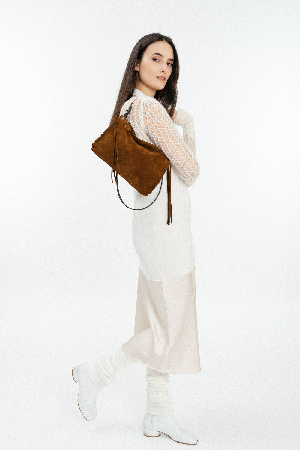 Woman standing in profile wearing a white outfit and boots, holding an INA KENT brown suede shoulder bag against a plain white background.