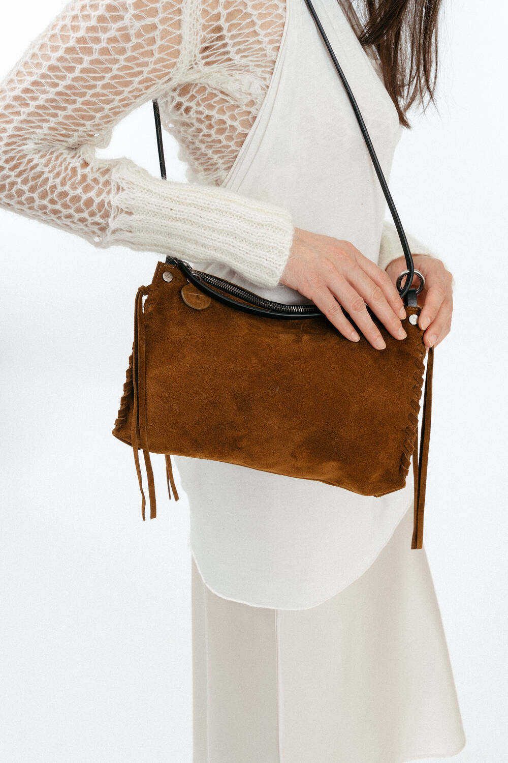 A woman wearing a white dress and knit top holds an INA KENT brown suede shoulder bag with a black strap and tassel details.