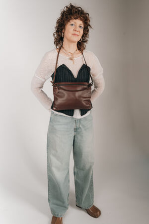 Das Model, gekleidet in Jeans, einen hellen Pullover mit einem dunkelblauen Korsett drüber, trägt eine INA KENT Umhängetasche MOONLIT ed.1 aus braunem Glattleder um den Hals, mit den Händen hinter dem Rücken verschränkt