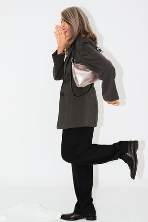 A woman with gray hair, wearing a black blazer and pants, black shoes, and holding an INA KENT metallic bag, stands with one leg raised, smiling and covering her face with one hand.