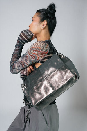 Person with a high bun hairstyle seen in profile, wearing a patterned, long-sleeve shirt and holding a large INA KENT bag over their shoulder.