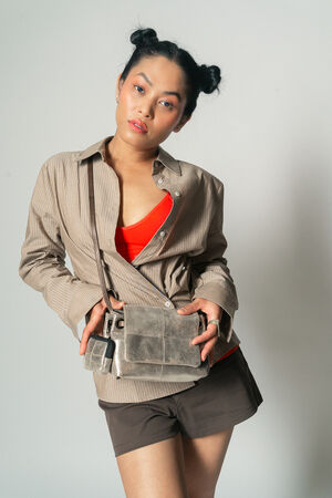 A person poses against a plain background wearing a brown shirt, red top, brown shorts, and a beige INA KENT crossbody bag. They have their hair styled in two buns.