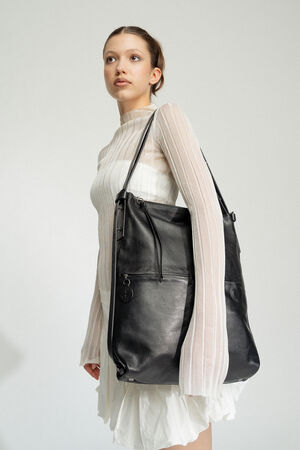 A young woman in a white sheer long-sleeve dress elegantly carries a large black INA KENT leather bag over her shoulder, standing against a plain background.