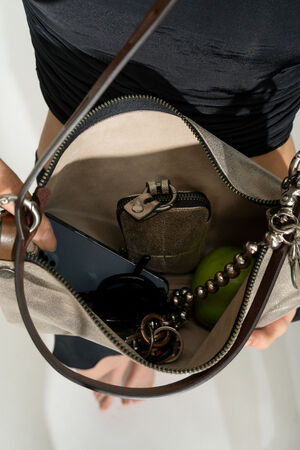 A close-up of an open INA KENT handbag containing a wallet, phone, keys, an apple, and a beaded bracelet. The person is holding the bag with one hand while wearing a black outfit.