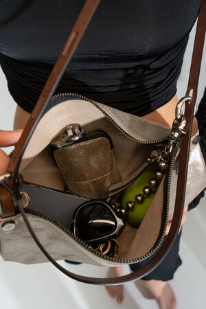 A person is holding an open purse revealing its contents: a smartphone, INA KENT sunglasses, a green apple, a small gold-colored bag, and decorative beads.