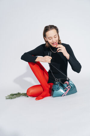 Woman in a black top and red tights sits on the floor and holds a candy cane in her hand. Next to her is the INA KENT ORNAMENT ed.2 bag in crackled dark teal leather, decorated with more candy canes and a small twig.