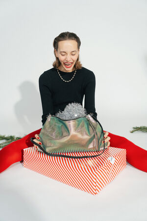 Woman in a black top and red tights sits behind a red and white striped gift package. On top is the INA KENT bag in two-tone leafy rose leather, filled with silver tinsel.