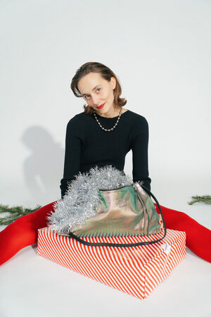 Woman in a black top and red tights laughs as she sits behind a red and white striped gift package. The INA KENT bag in two-tone leafy rose leather, filled with silver tinsel, lies on top.