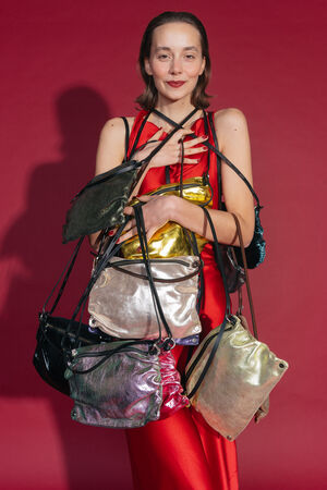 A smiling person in a red silk dress holds a variety of MOONLIT ed.1 bags in different metallic colors, including gold, silver, black, green and pink. The bags with their elegant straps are artfully draped around the person against a festive red background.