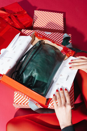 Person in red tights and black top opens a gift with red and white striped paper. In a red gift box lies the MOONLIT ed.1 bag in the color Patina Green. The metallic green bag is elegantly wrapped in printed tissue paper, surrounded by other festively wrapped gifts in red and white.