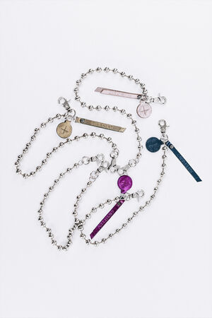 The picture shows several silver-colored ball chains. Each chain is combined with a colored pendant, including round plaques and narrow leather strips in various colors such as gold, pink, purple and blue. Some of the pendants are embossed with “INA KENT”. The chains lie loose on a white surface.