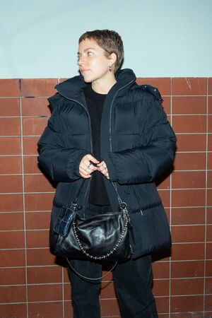 A female-presenting person looks to the side while holding a black bag named "ORNAMENT ed.2" in front of a wall with red tiles.