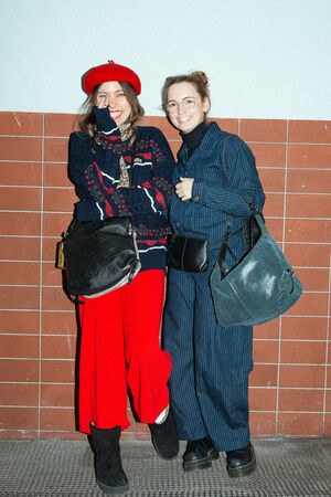 Two female figures are standing next to each other. The person on the left is wearing a red hat, a patterned sweater, red trousers and black boots. The “AD LIB ed.1” bag in pure black with a gold-colored tag hangs over her shoulder. The person on the right is wearing a dark blue suit with pinstripes, black shoes and two bags: an “X.MOG ed.2” in pure black as a hip bag and a “DELA YIN ed.1” over the shoulder.