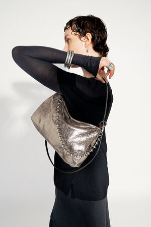 Model with INA KENT ORNAMENT ed.2 metallic bright anthra (side view with ball chain): The model wears the metallic anthracite-colored bag over her shoulder. The bag features a silver ball chain as a decorative element that runs along the front. The metallic shine of the bag contrasts with the model's black clothing.