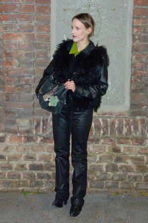  A female figure stands in front of a brick wall wearing a black leather jacket and holding the DELA EDEN bag in shimmering film noir.