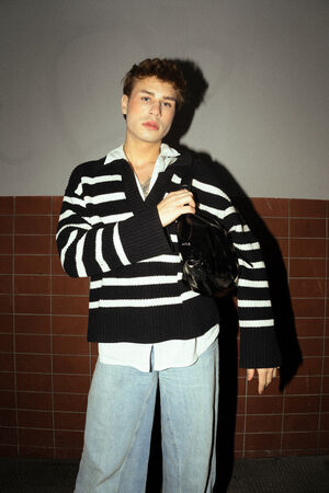 A male figure is wearing a black and white striped sweater and blue jeans. The “DINKUM ed.2” bag in patent black hangs over his shoulder.