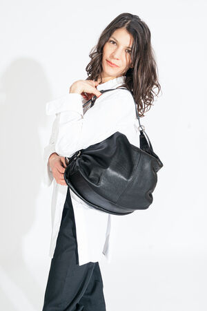 A person in a white shirt and dark pants confidently holds an INA KENT large black leather bag against a pristine white background.