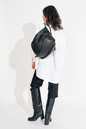 A person with long hair, dressed in a white shirt, black pants, and knee-high boots, carries an elegant INA KENT bag against a plain backdrop.