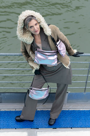 Die Frau lehnt am Geländer eines Stegs mit einer metallic-lavender-rose INA KENT Tasche als Crossbody-Bag und einer weiteren kleineren Tasche an der Schulter. Die Perlenkette am Riemen betont den eleganten Stil.